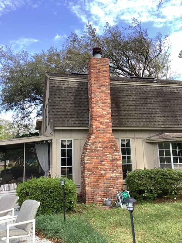 Tall chimney on side of barn shaped house with lots of tall windows, carport, chairs to the left and trees and blue cloudy sky in the background. Time for a virtual inspection.