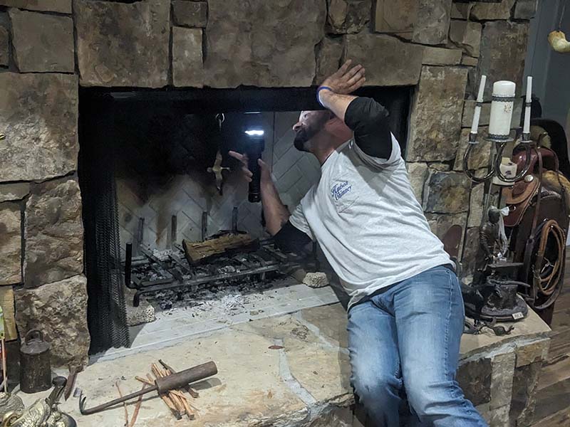 Technician performing an inspection sitting on stone hearth using a flashlight.  There is a saddle sitting on a stand to the right behind him.